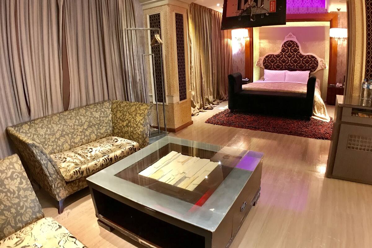 A hotel room with a sofa, a glass coffee table, a bed with a patterned headboard, and a TV mounted on the wall.