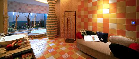 A modern hotel room with a checkered floor, a round pillar, a sofa with cushions, a small table with a vase, and a window with a view of trees.