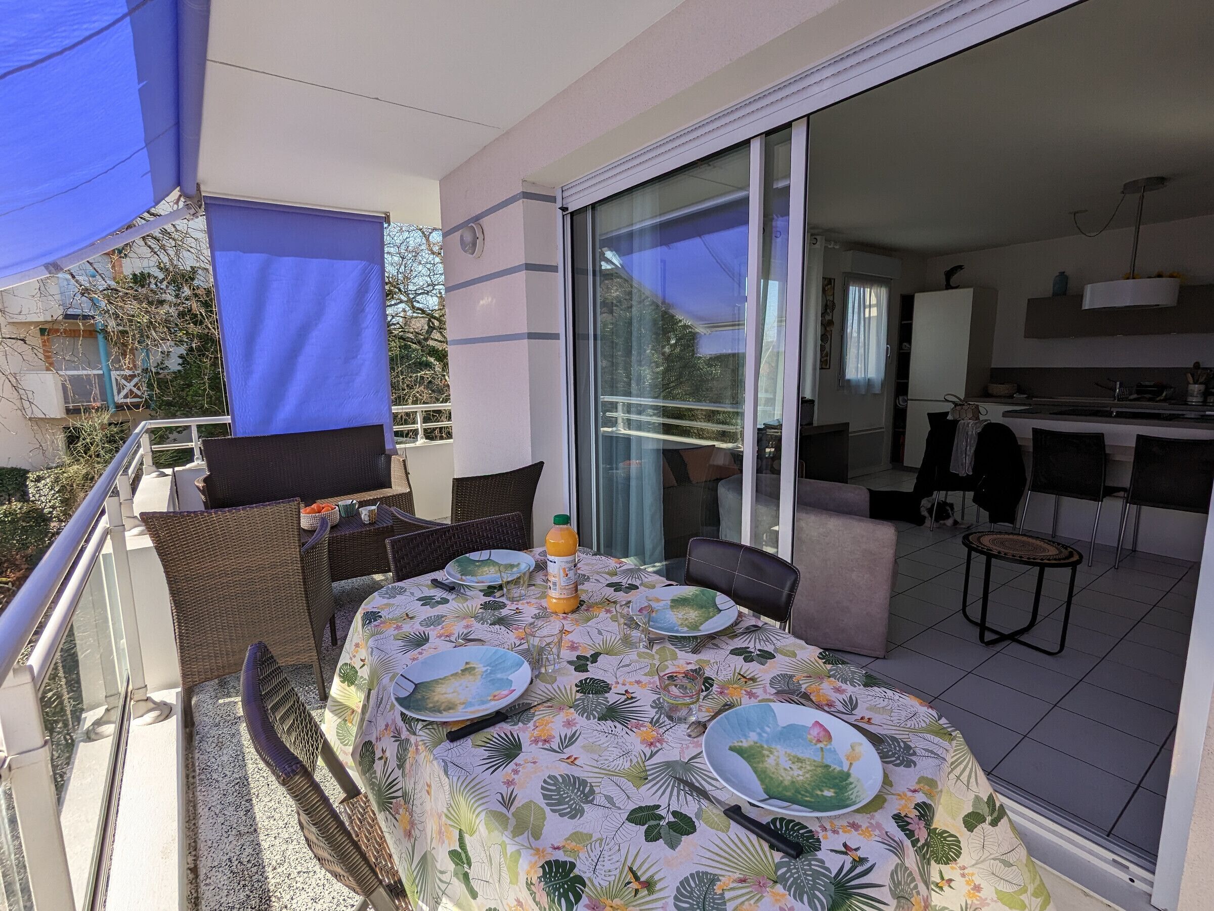 An outdoor dining area with a table set for two, a blue umbrella, and a view into the kitchen.