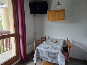 Una habitación con una mesa, sillas, un televisor montado en la pared y un ventanal con cortinas.