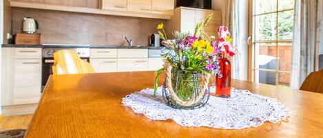 Ein Holztisch mit einer Spitzendecke, eine Vase mit Blumen und eine rote Flasche. Der Raum verfügt über Holzschränke, eine Küchenzeile und ein Fenster mit Vorhängen.