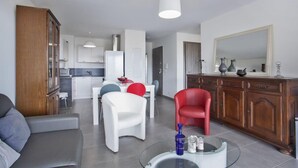 A modern living room with a sofa, a dining table with chairs, a red chair, a glass coffee table, and a kitchen area in the background. - Casa San Ghjisé (Bastia)