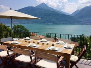 Mesa de madera preparada para comer, con platos y vasos blancos, un paraguas color beige, y vista a un lago y montañas.