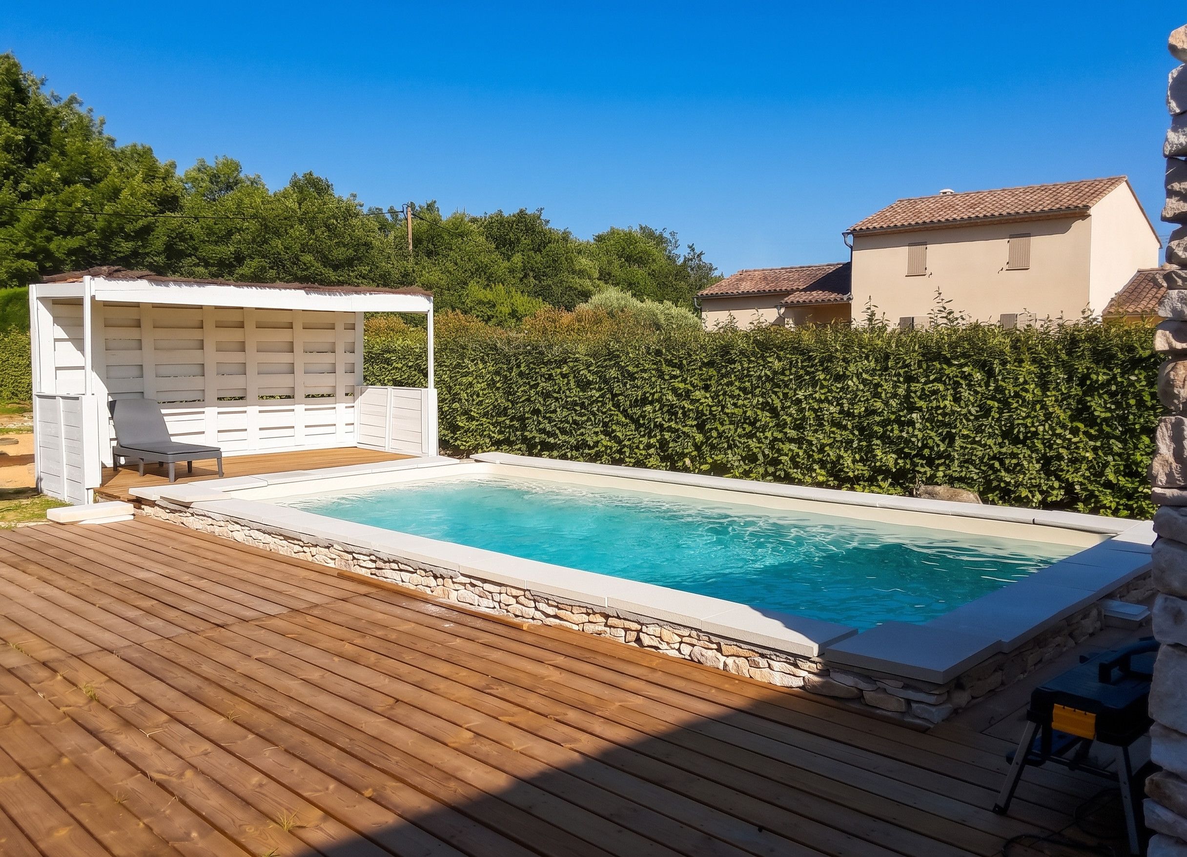 Ein Schwimmbad mit Holzterrasse, überdachter Sitzecke und einer Hecke im Hintergrund.