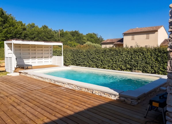 Una piscina con muelle de madera, zona de estar techada y seto al fondo.