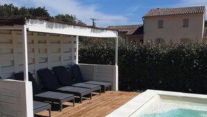 Una terraza de madera con piscina, un espacio de descanso cubierto con cojines y un seto al fondo.