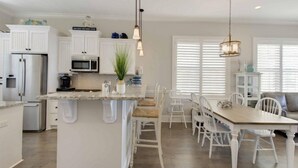 A spacious kitchen with white cabinetry, a central island, and a dining area with white chairs. - Ocean Cottage 39 Halekulani The Cottages at Jekyll Island Oceanfront (Jekyll Island)