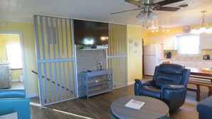 Un espace de vie compact comprenant un canapé, un fauteuil, une petite table, un ventilateur de plafond, un téléviseur et un coin cuisine avec un réfrigérateur et un évier.