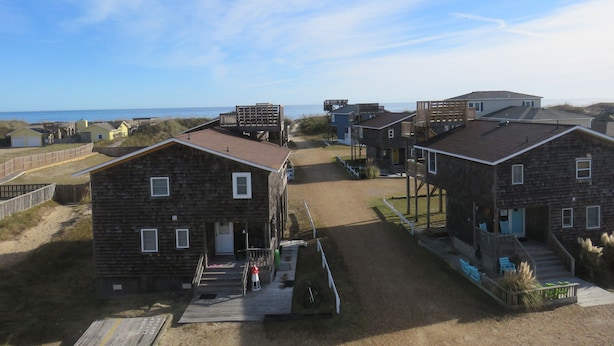 Une zone résidentielle côtière avec de nombreuses maisons en bois, dont certaines sont dotées de terrasses surélevées, et une plage de sable en arrière-plan.