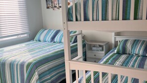 A room with a bunk bed and a single bed, both with striped bedding. There is a small bedside table with a lamp and a drawer. - Great Condo With Ocean Views!! (Brigantine)