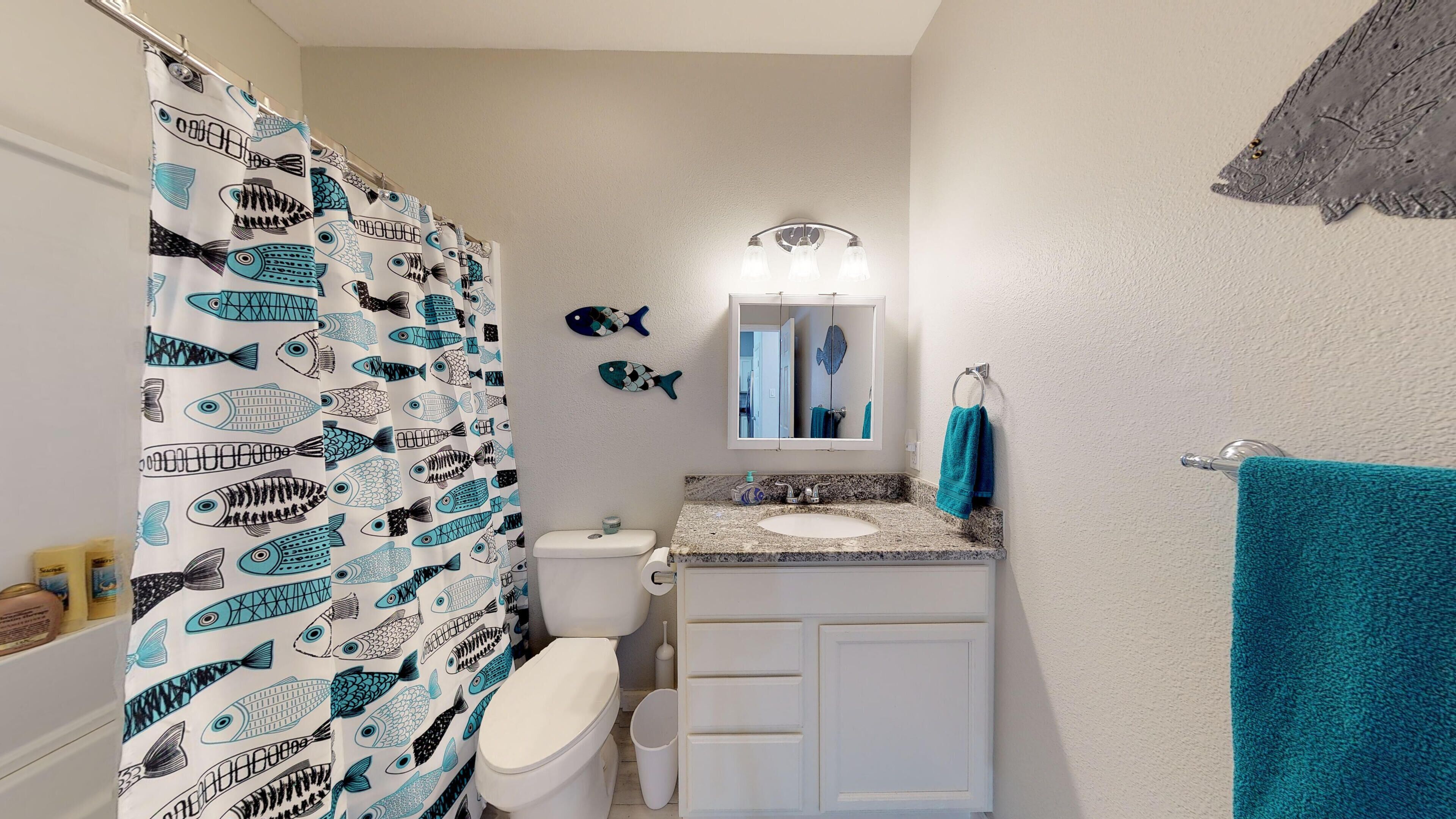 A bathroom with a shower curtain featuring fish patterns, a toilet, a sink with a mirror above it, and a towel rack with a blue towel.