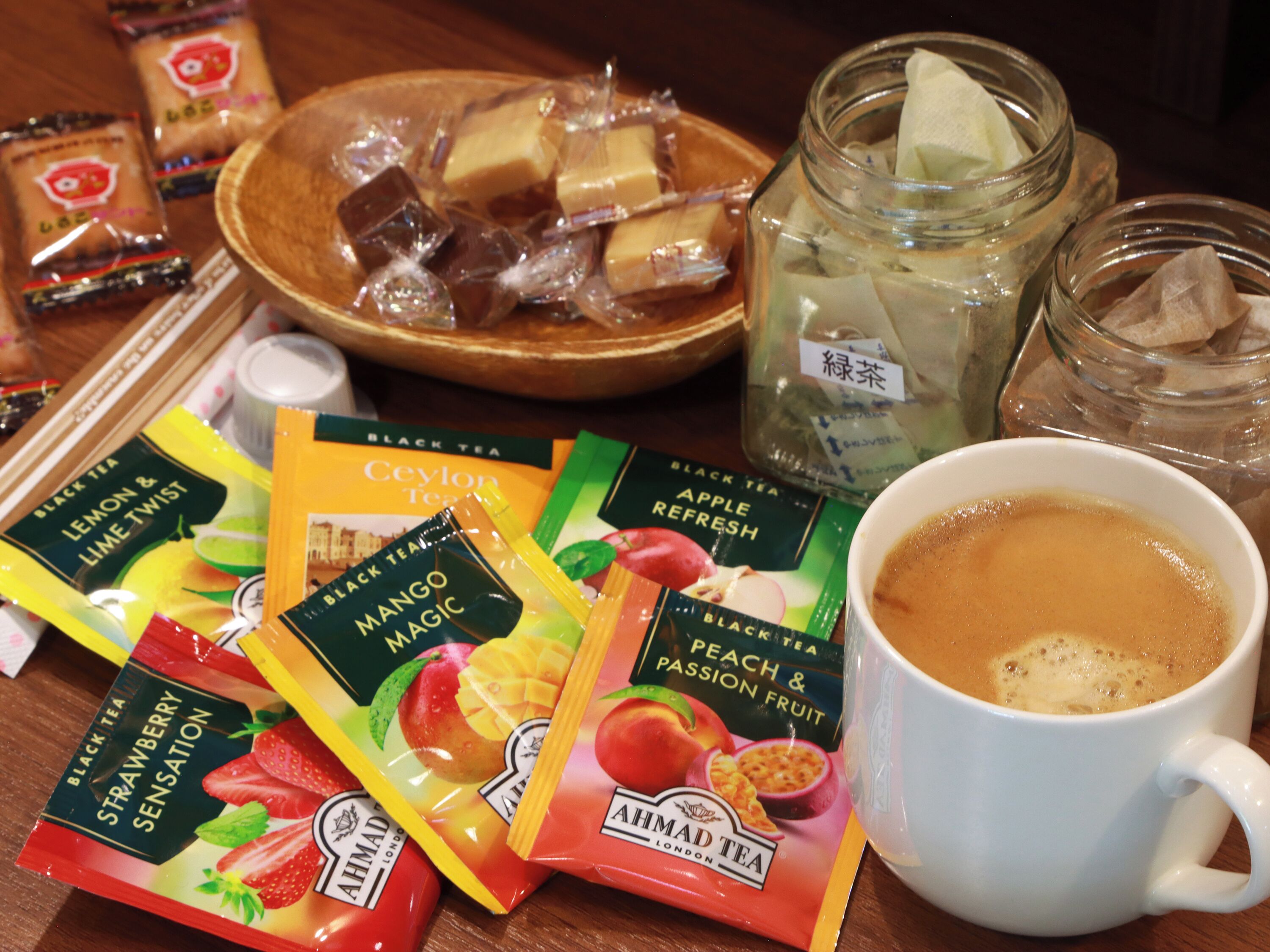 A variety of tea packages, a bowl of assorted chocolates, a mug of coffee, and a jar with a tea bag.