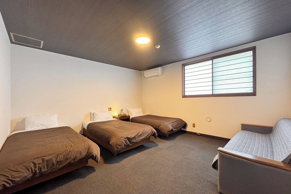 A room with two single beds, a sofa, a window with blinds, and a ceiling light.