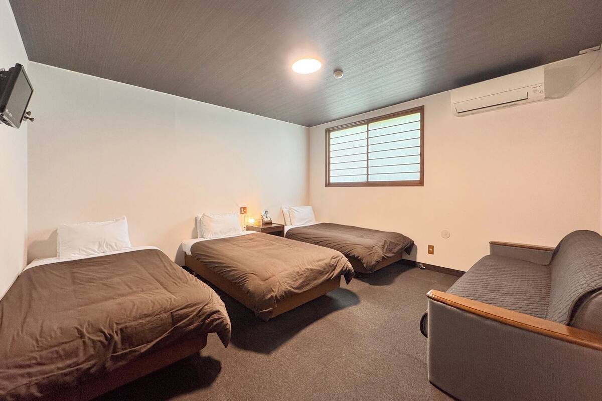A room with two beds, a sofa, a window with blinds, a ceiling light, and an air conditioning unit.