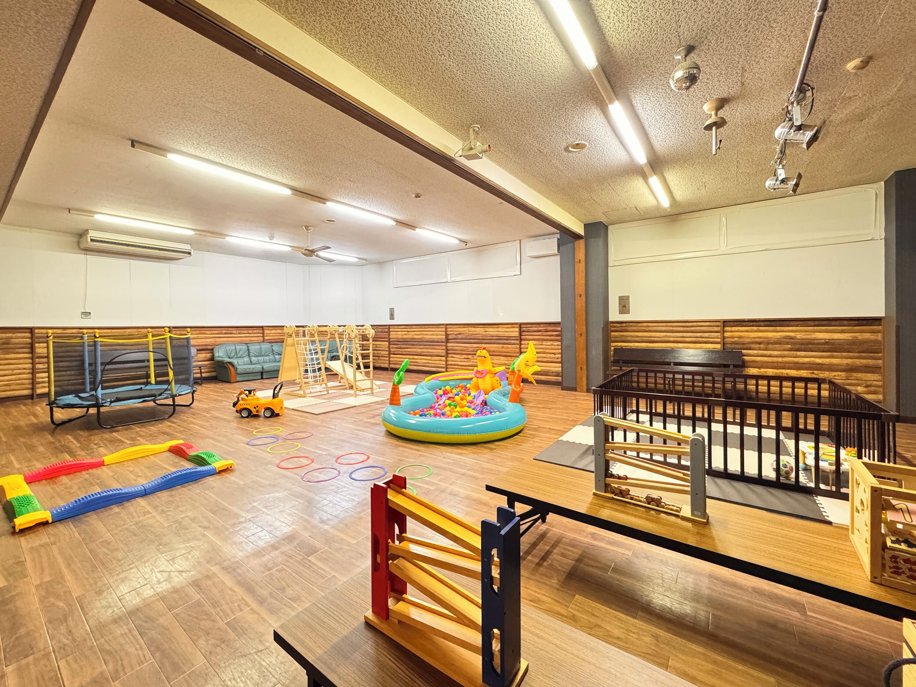 A spacious indoor play area with a wooden floor, a swing set, a ball pit, and a small playhouse.