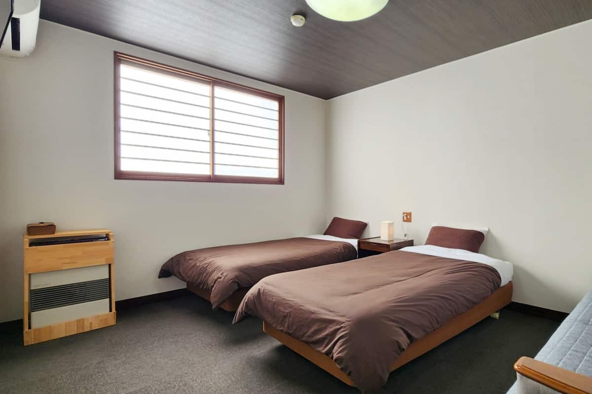A room with two beds, a wooden nightstand, a small table, and a window with blinds.