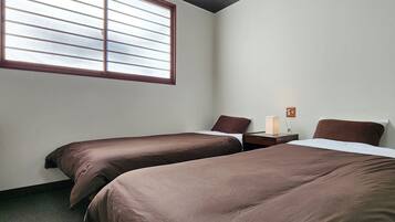 A hotel room with two single beds, a nightstand with a lamp, and a window with blinds.