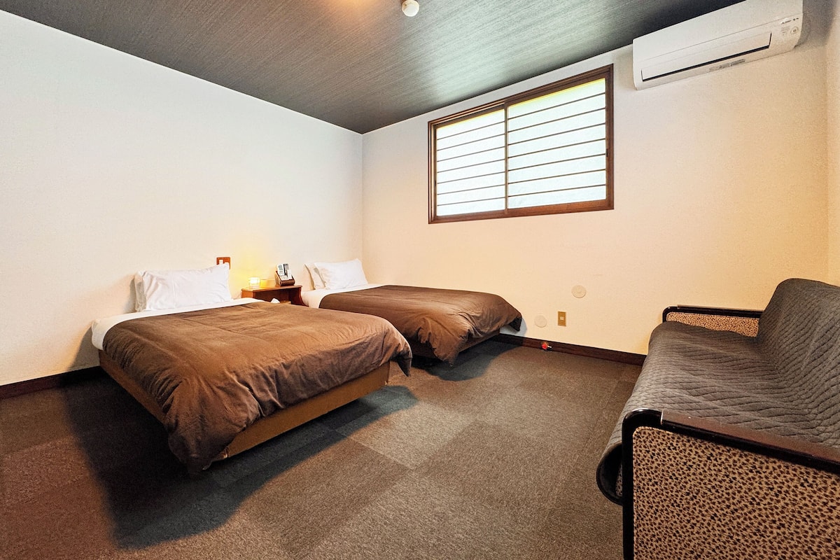 A hotel room with two beds, a single chair, a window with blinds, and an air conditioning unit.