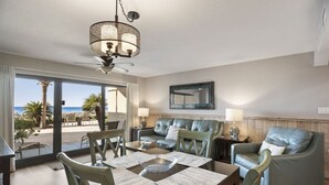 Uma sala de jantar com uma mesa posta para quatro pessoas, um sofá e uma cadeira. Há um ventilador de teto sobre a mesa e vista para a praia através das portas de vidro.