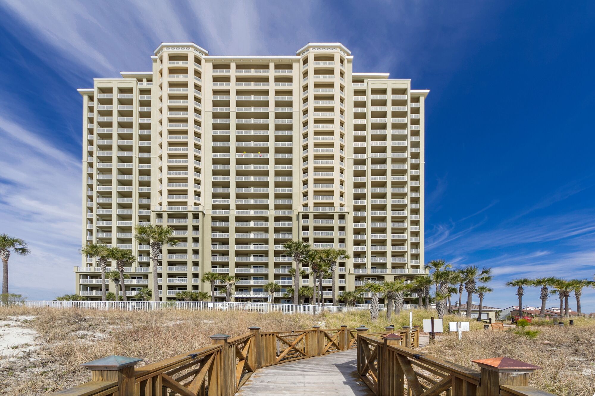 A high-rise hotel with multiple balconies, a wooden boardwalk leading to the entrance, and palm trees in the foreground.