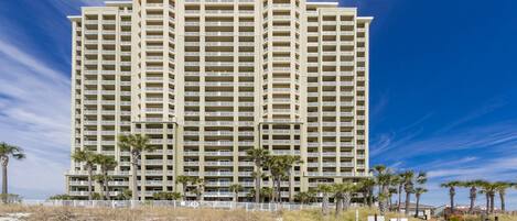 A high-rise hotel with multiple balconies, a wooden boardwalk leading to the entrance, and palm trees in the foreground.