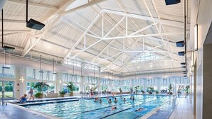 Une piscine intérieure dotée de plusieurs couloirs, de grandes fenêtres et d’un plafond haut orné de motifs géométriques.