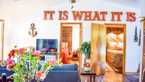 Una sala de estar con ventilador de techo, televisor, sofá y una mesa de centro con flores. Se ve una cama con cabecera de madera al fondo.