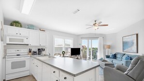 A modern kitchen with white cabinetry, a built-in microwave, and a large island. There is a ceiling fan, a dining area with a table and chairs, and a living room with a sofa and a recliner.