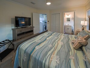 Ein Schlafzimmer mit einem Bett, einem Fernseher auf einer Holzkommode, einer Waschmaschine und einem durch eine offene Tür einsehbaren Badezimmer.