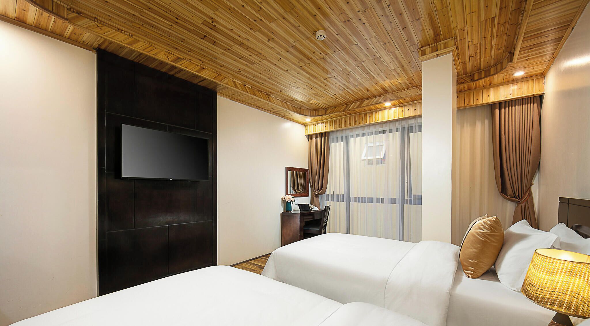 Ein Hotelzimmer mit Holzboden, einem Flachbildfernseher an einer dunklen Wandverkleidung, einem Schreibtisch mit einem Laptop und einem Bett mit weißer Bettwäsche und einem goldenen Kissen.