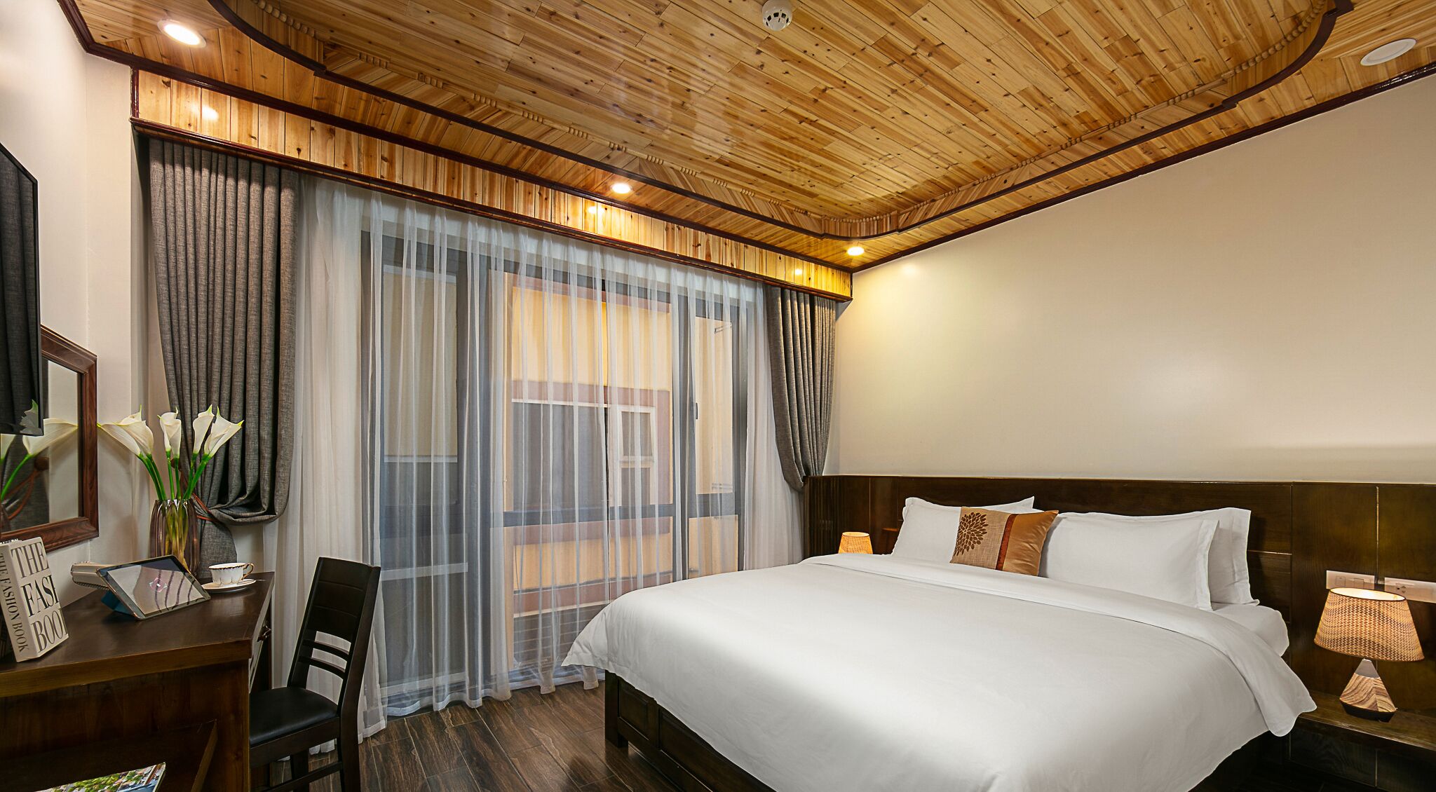 A hotel room with a wooden ceiling, a large bed with white linens, a desk with a chair, and a large window with sheer curtains.