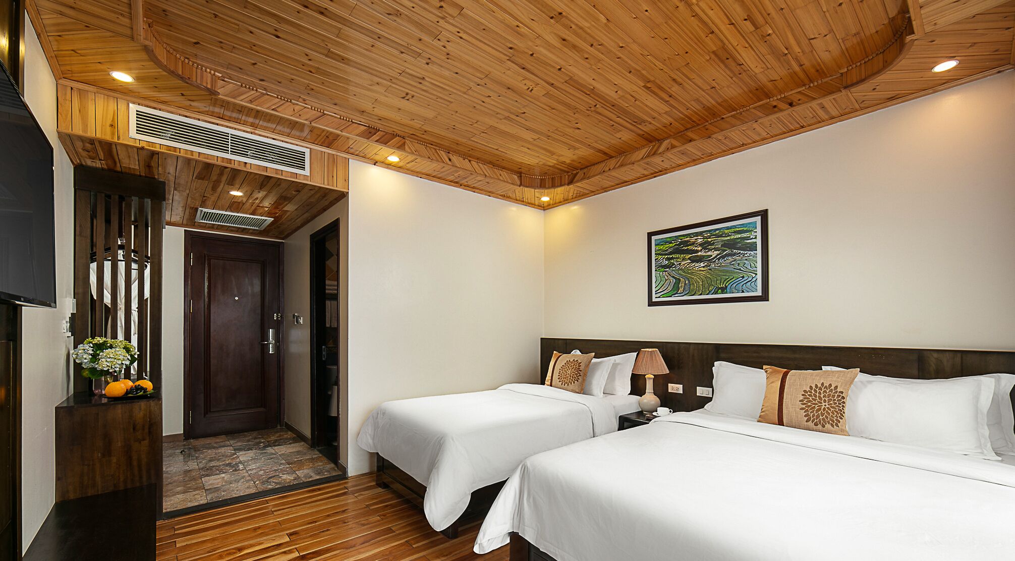 Een hotelkamer met twee bedden, houten vloer, houten plafond, een schilderij aan de muur en een tafeltje met een lamp.