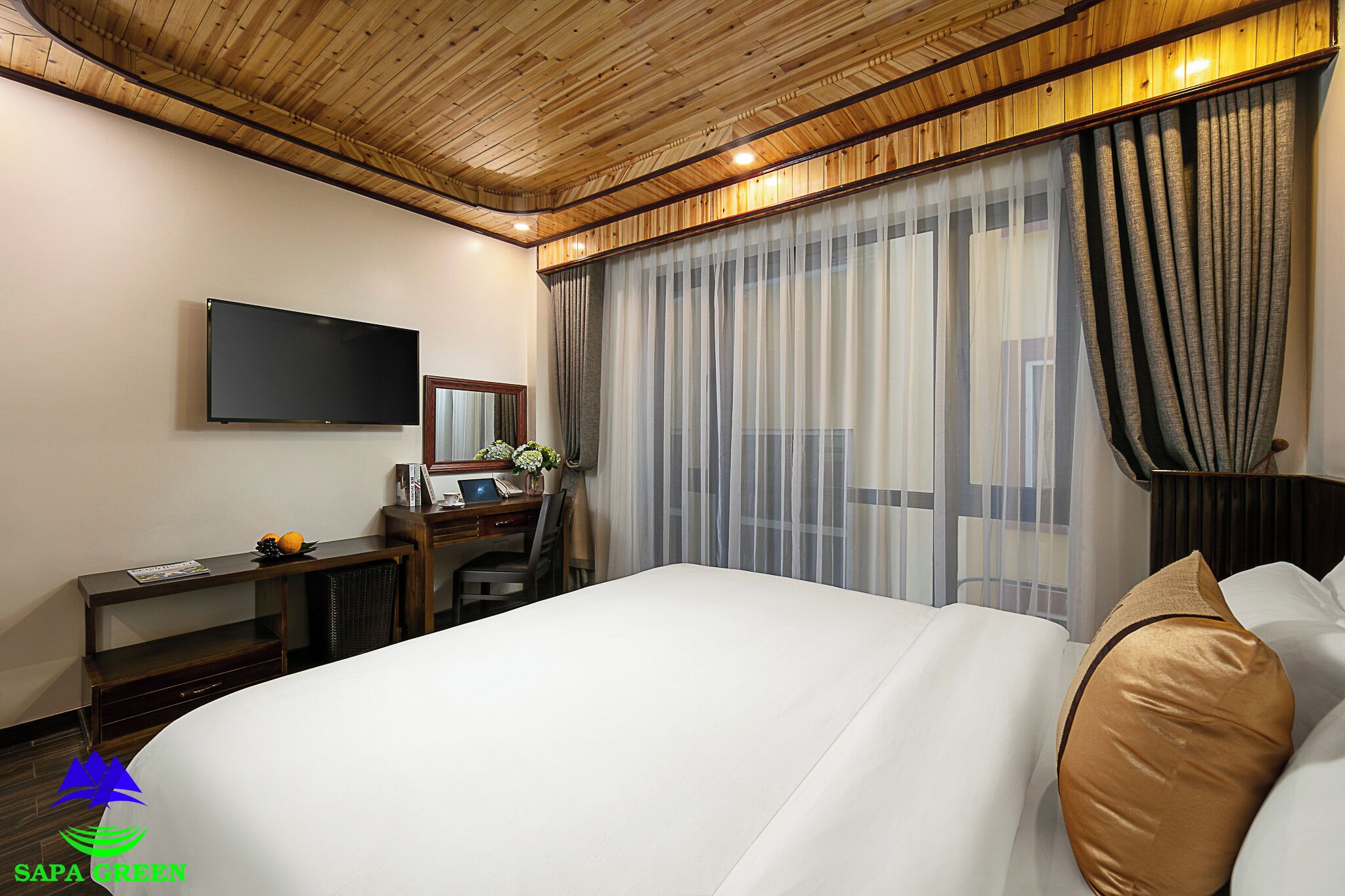 Quarto de hotel com uma cama grande, uma escrivaninha com computador, uma televisão de tela plana e uma janela ampla com cortinas.