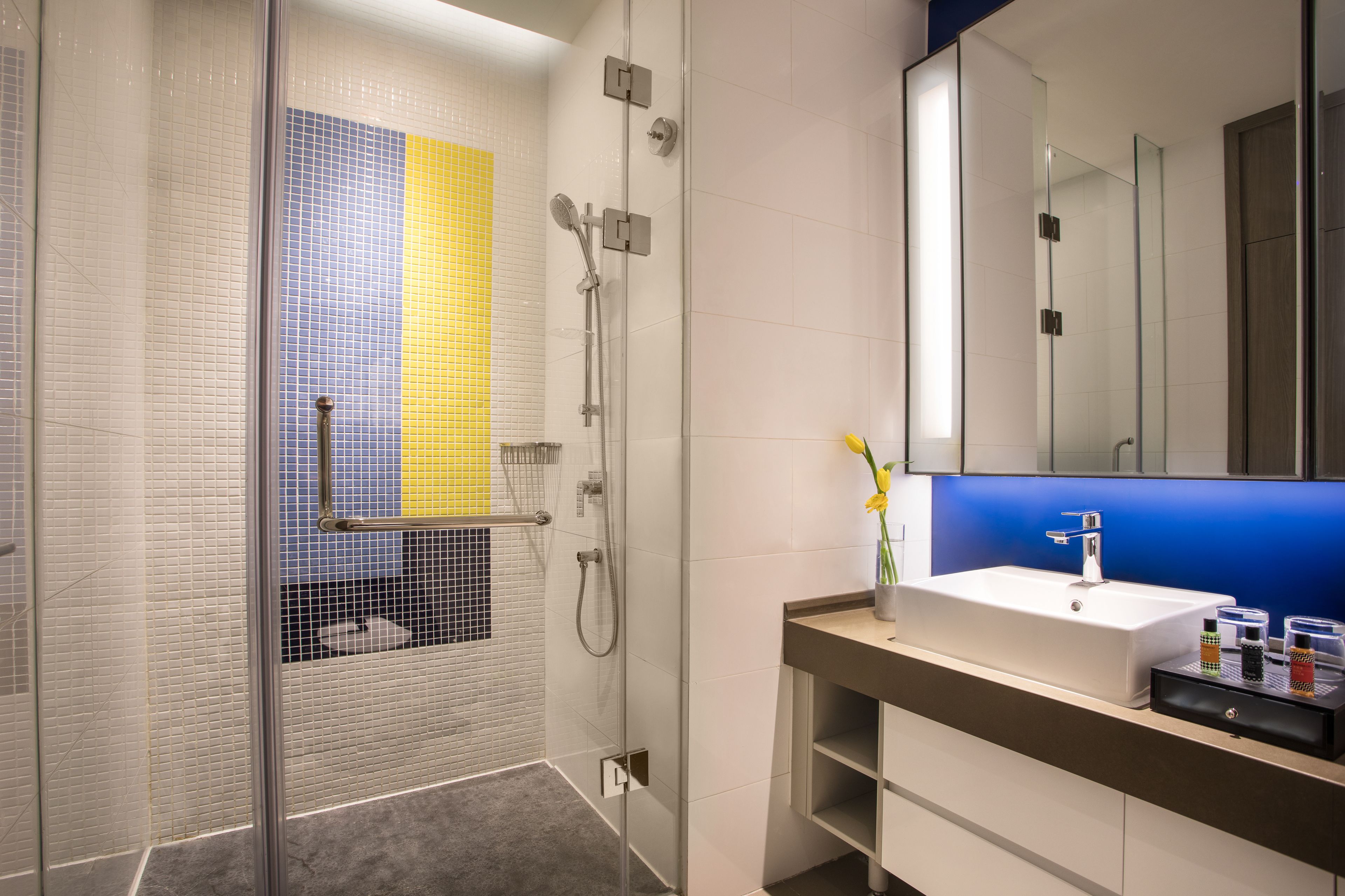 A modern bathroom with a glass-enclosed shower, a white sink with a blue-lit mirror, and a tiled wall with yellow and blue accents.