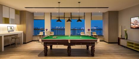 Una habitación moderna con mesa de billar, sillas, escritorio y un amplio ventanal con vista a la ciudad.