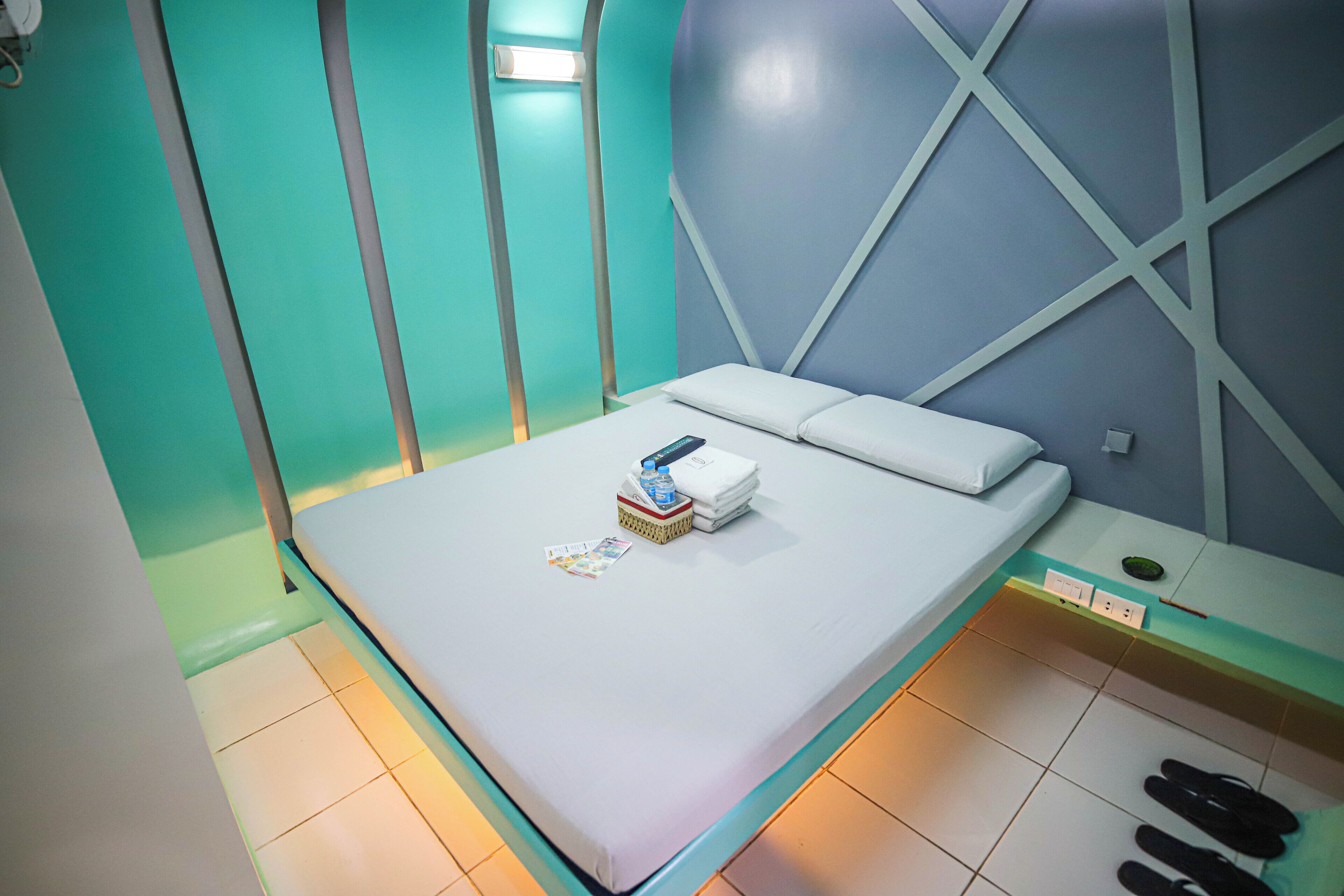 A neatly made bed with a white mattress and pillows, a small bedside table with a lamp, and a pair of slippers on the floor.