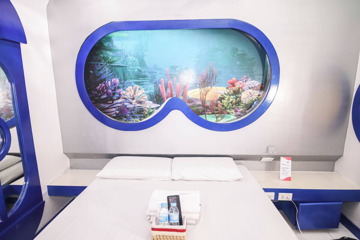 A room with a large aquarium window, a bed, a bedside table with a remote control, a water bottle, and a magazine.