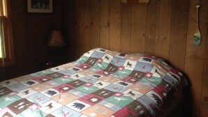 Een slaapkamer met een houten wand, een bed met een patchworkdeken, een lamp en een lijst met een afbeelding aan de muur.