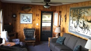 Interior acogedor de una cabaña con paredes y detalles de madera, un sofá, una silla, una lámpara y un cuadro en la pared.
