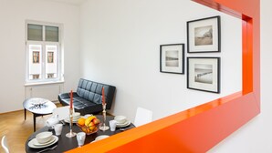 Ein modernes Wohnzimmer mit einer schwarzen Ledersofa, einem Esstisch, der mit weißen Tellern, Tassen und einer Obstschale gedeckt ist. An der Wand hängen gerahmte Bilder und es gibt ein Fenster mit Jalousien.