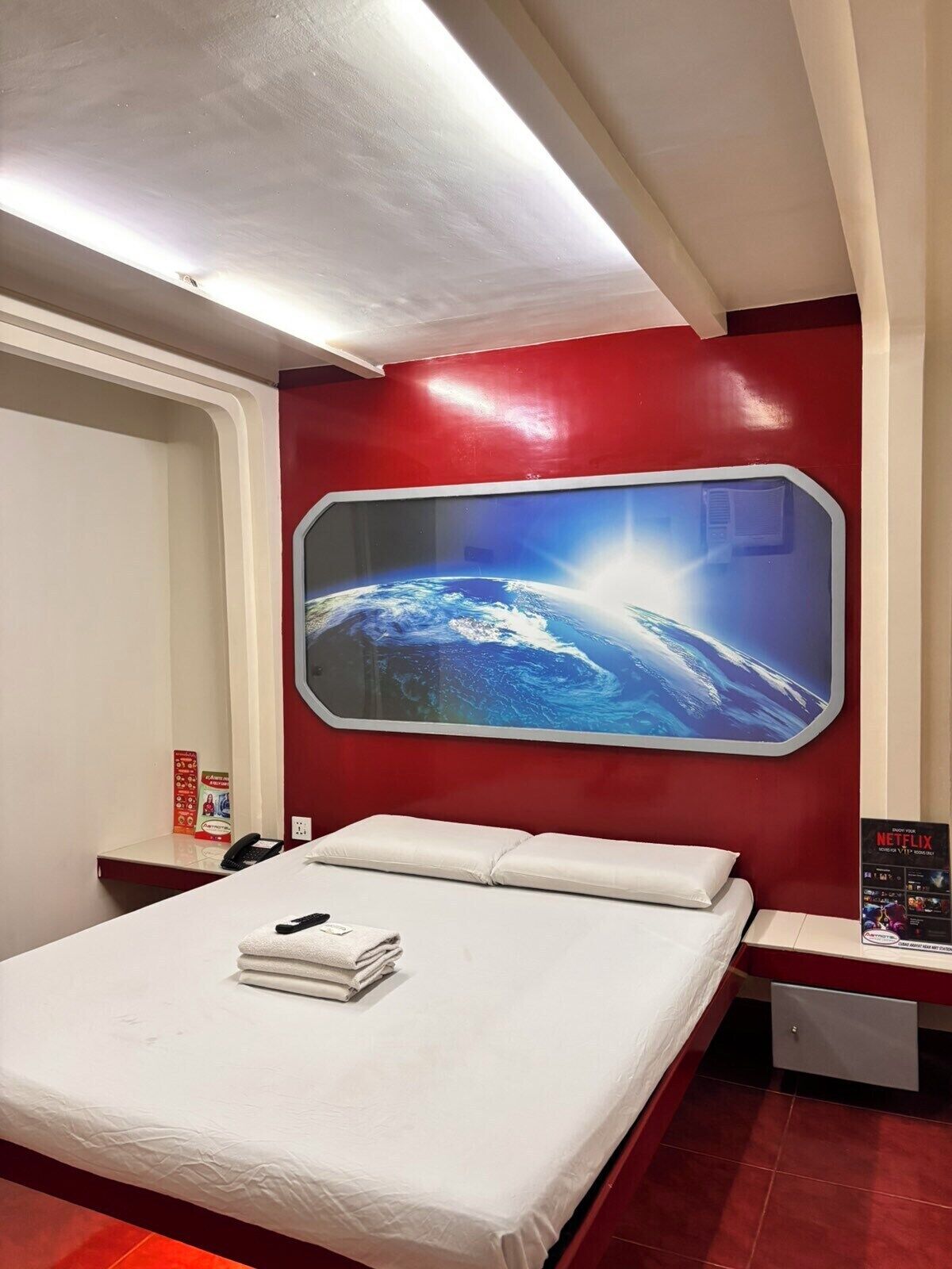A hotel room with a large window, a bed with white linens, a red wall, and a poster on the wall.