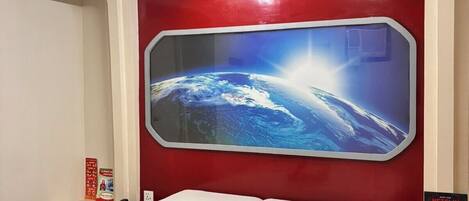 A hotel room with a large window, a bed with white linens, a red wall, and a poster on the wall.