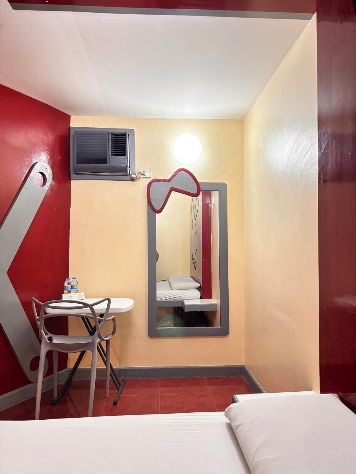 Camera d'albergo compatta con un letto, un piccolo tavolo con sedie, uno specchio e un condizionatore d'aria.