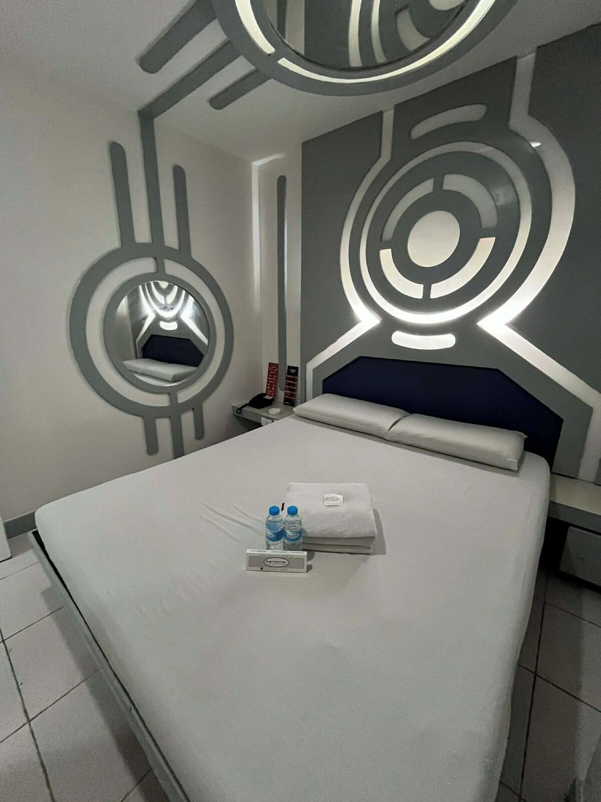 A modern hotel room with a bed, a nightstand, a mirror, and a wall with geometric light fixtures.