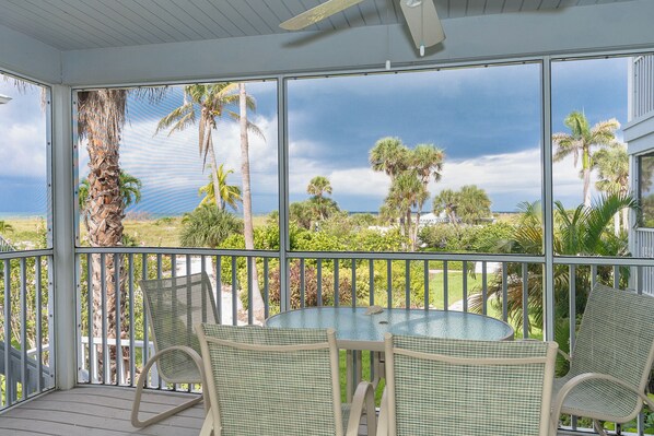 A covered outdoor area with a table and chairs, large windows offering a view of palm trees and greenery.