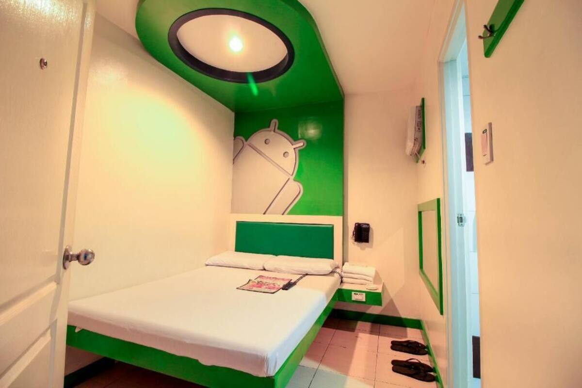 Une petite chambre avec un lit simple, un mur d’accent vert, un luminaire au plafond, un téléphone fixé au mur et une porte dotée d’un cadre vert.