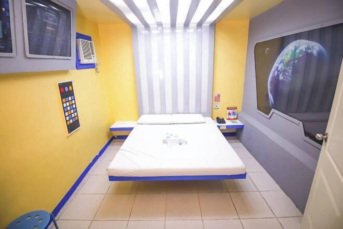 Une petite pièce, bien éclairée, avec un lit simple, une fenêtre donnant sur la Terre, un panneau de contrôle et une petite table avec une chaise.