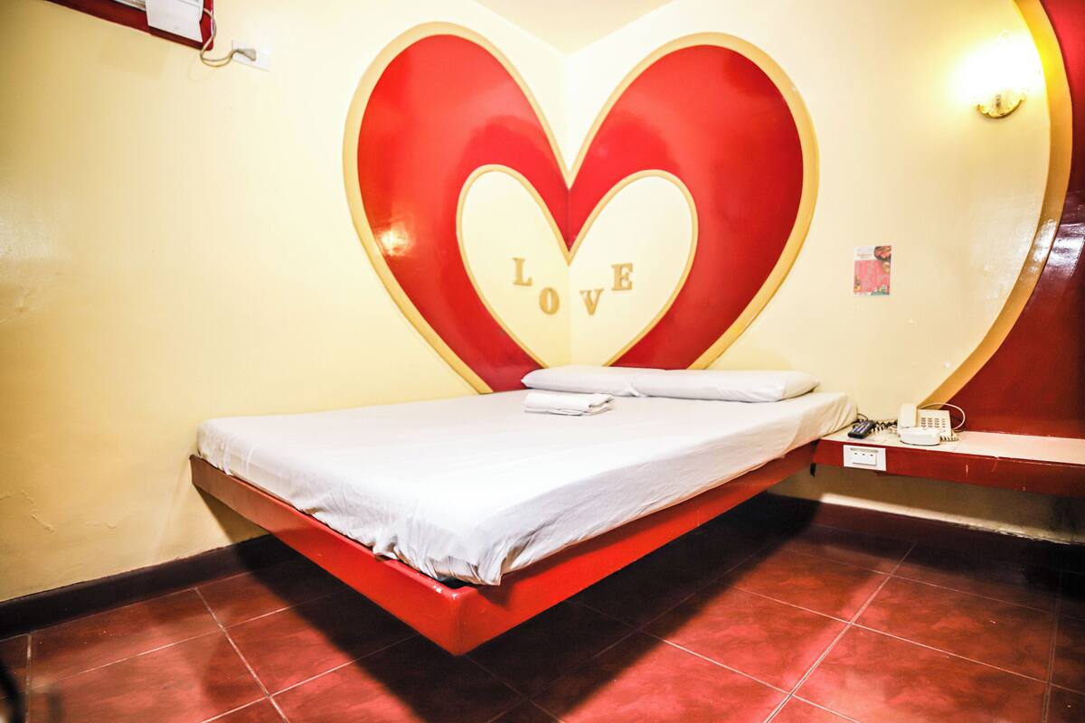 Un lit avec un cadre rouge et une grande décoration murale en forme de cœur, le mot « AMOUR » étant inscrit au centre.