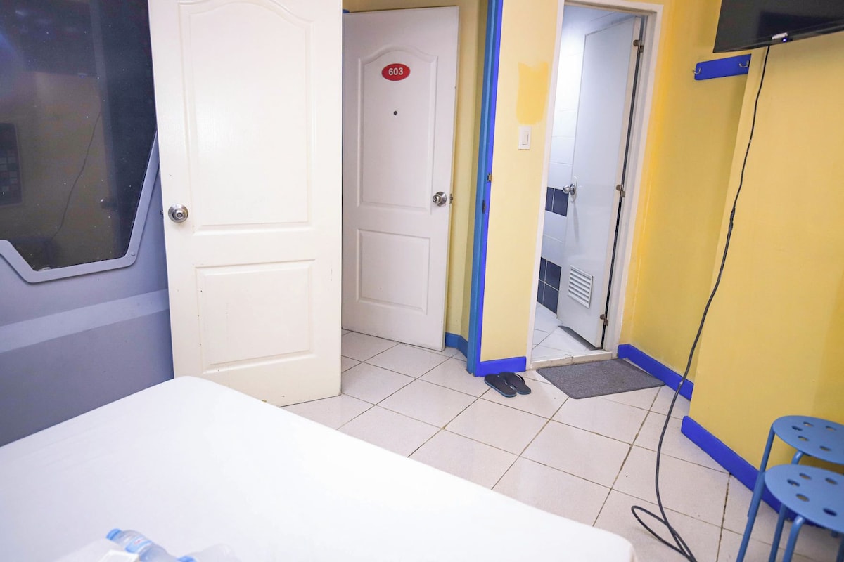 Habitación de hotel con cama, puerta numerada, baño con ducha y televisor montado en la pared.