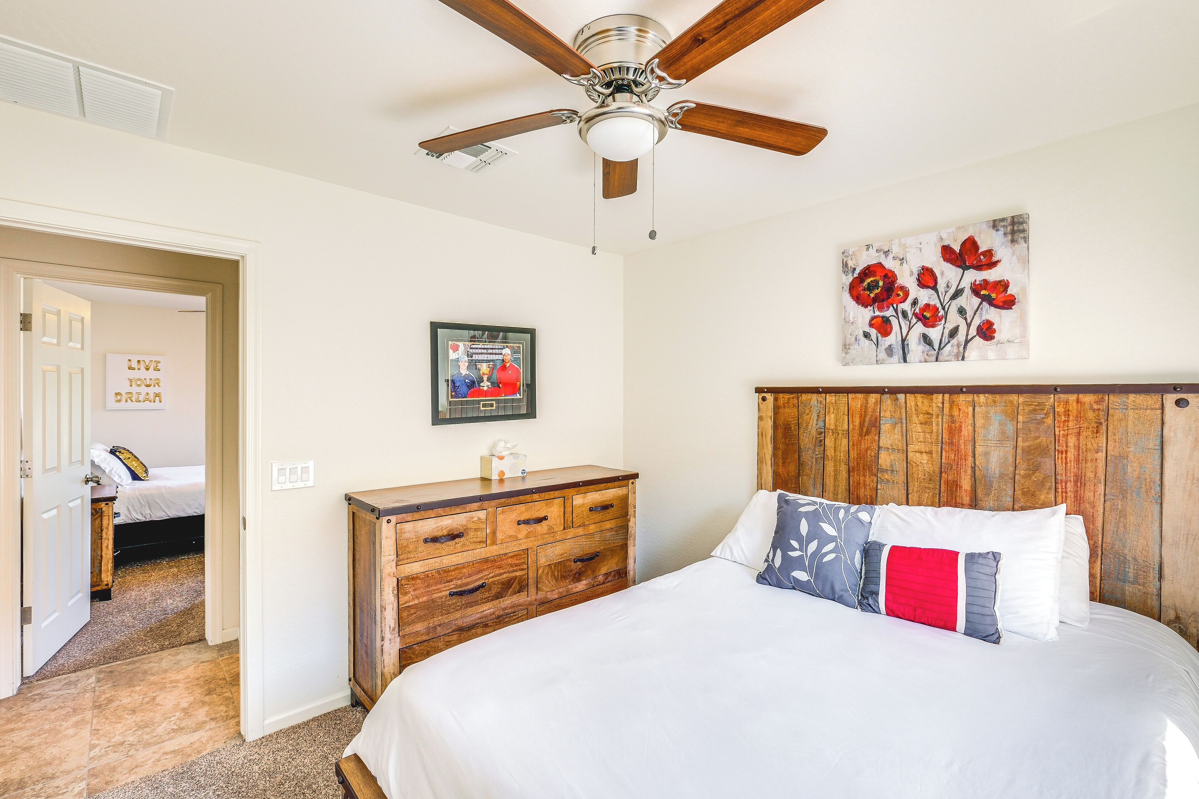 A bedroom with a wooden headboard, a ceiling fan, a bed with white bedding, a wooden dresser, and a painting of red flowers.